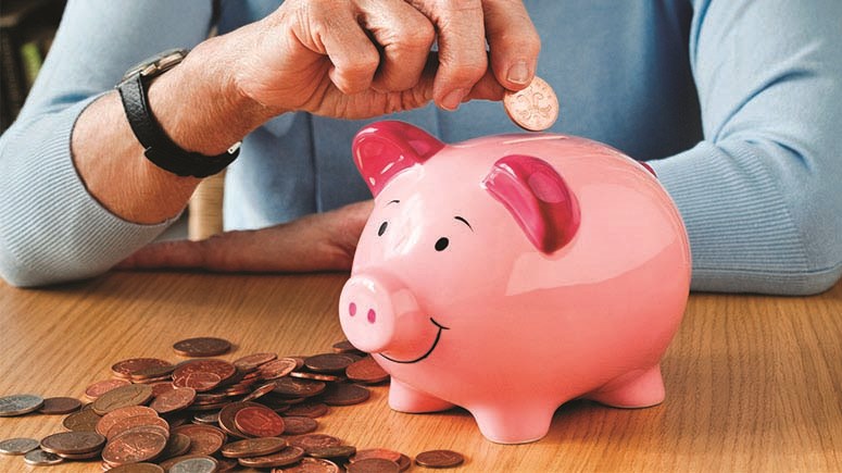 Close up on older person's hand putting a coin in to a piggy bank, and a stack of coins next to it